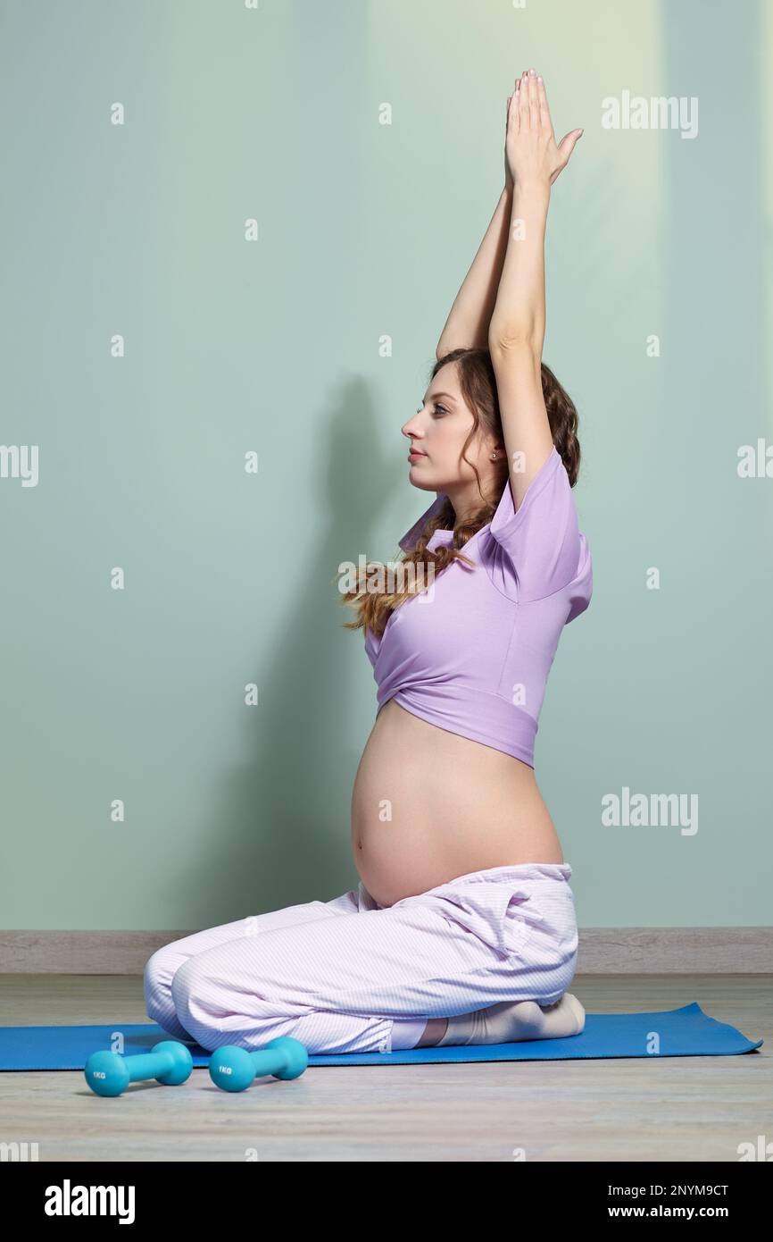 Una donna incinta con lunghi capelli castani siede a gambe incrociate, meditando e praticando yoga per il fitness fisico e il benessere durante la gravidanza. Mani in su. Foto Stock