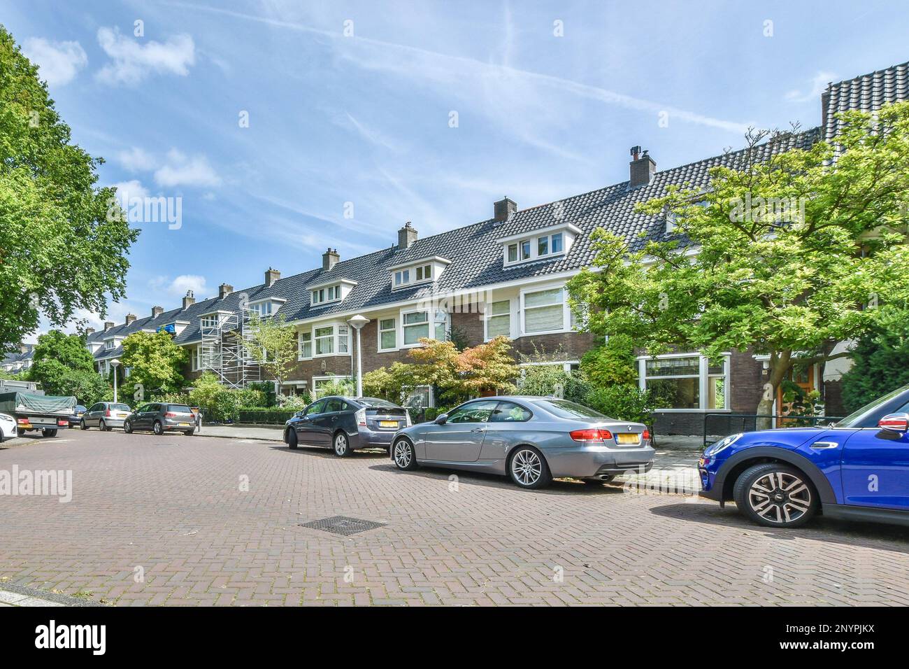 Amsterdam, Paesi Bassi - 10 aprile 2021: Alcune auto parcheggiate sul lato della strada di fronte a una fila di case con cielo blu e nuvole bianche Foto Stock