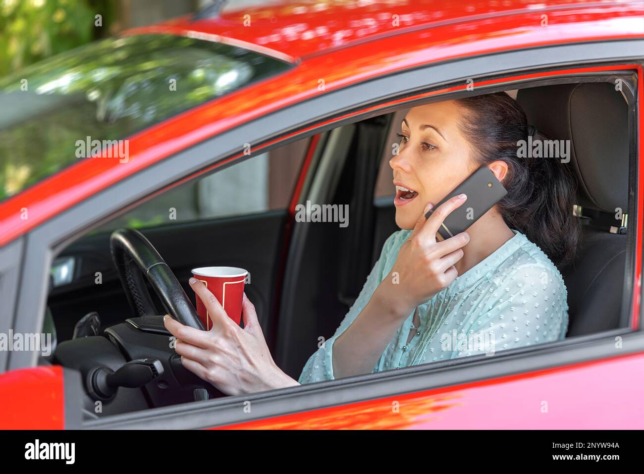 Una giovane donna conducente in auto con una tazza di caffè in mano parla al telefono. Foto Stock