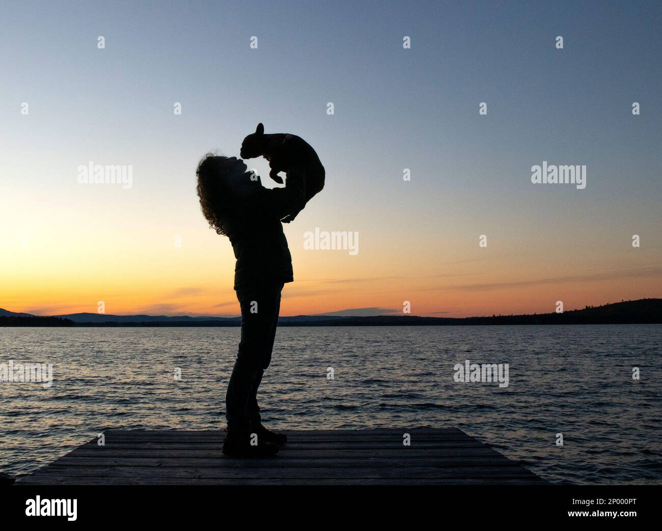 Donna con silhouette che tiene il bulldog francese sopra la testa dando bacio mentre si trova sul molo di fronte al tramonto sulle montagne nel nord del Maine boschi. Foto Stock