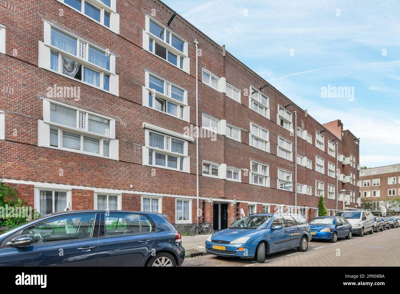 Amsterdam, Paesi Bassi - 10 aprile 2021: Alcune auto parcheggiate sul lato di una strada di fronte ad un edificio in mattoni con finestre bianche e porte chiuse Foto Stock