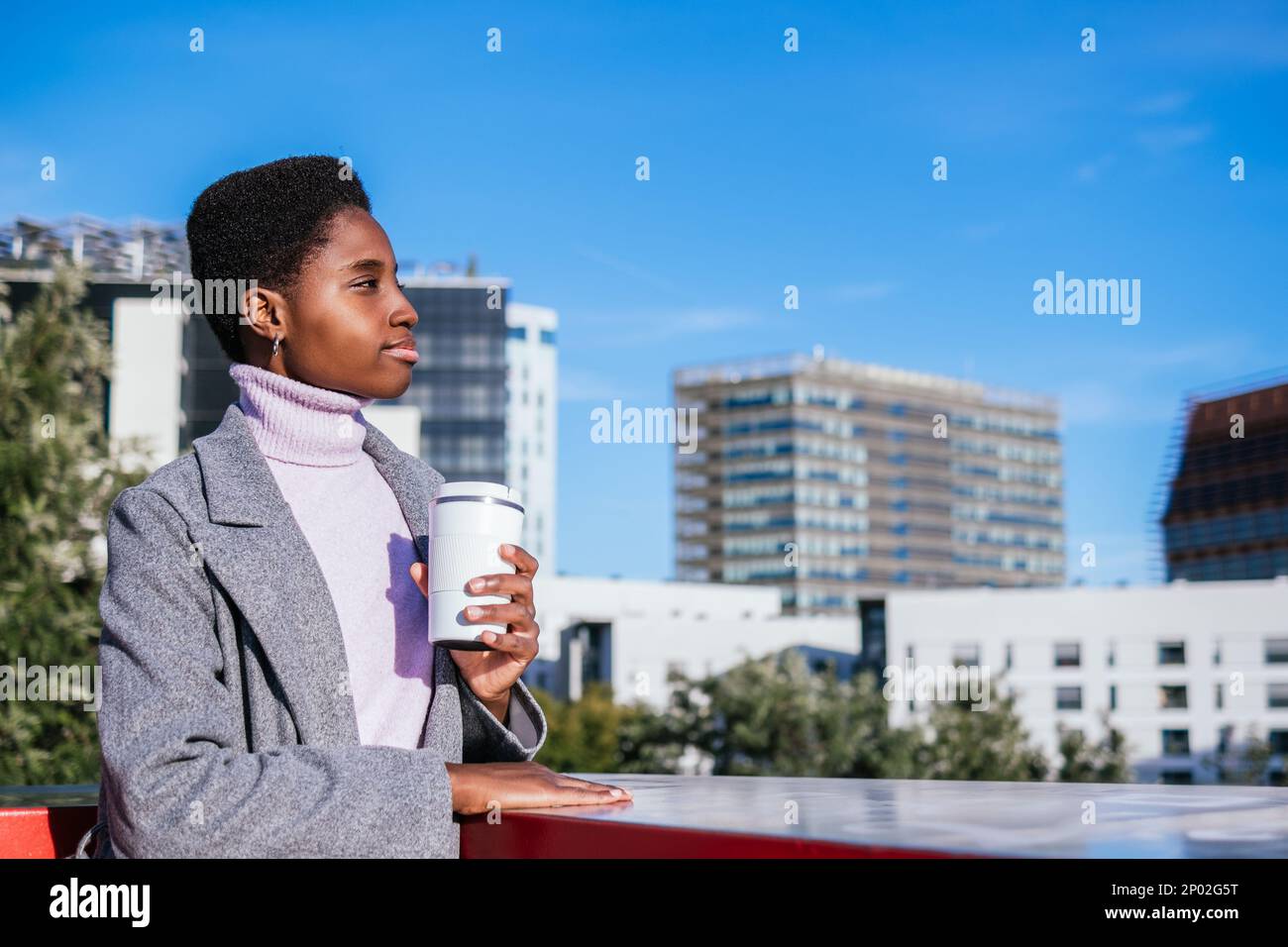 Ragazza afroamericana con una tazza di caffè ecocompatibile che guarda lontano e che ammira la strada della città moderna di giorno Foto Stock