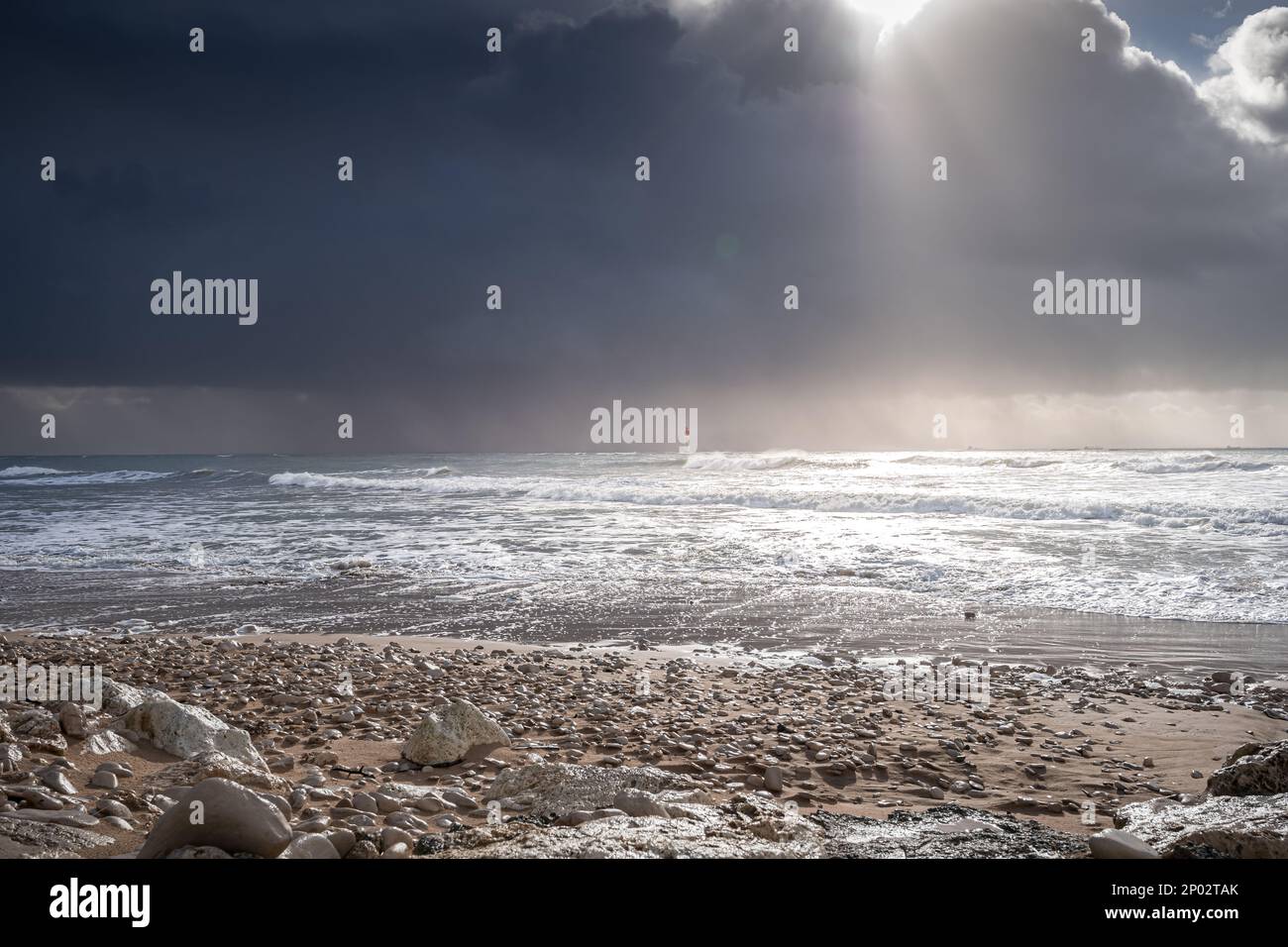 Spiaggia di ciottoli vicino all'oceano a Rivedoux, isola di Re, Francia. il sole splende tra le nubi grigie. Il faro di chauveau è in mare aperto Foto Stock