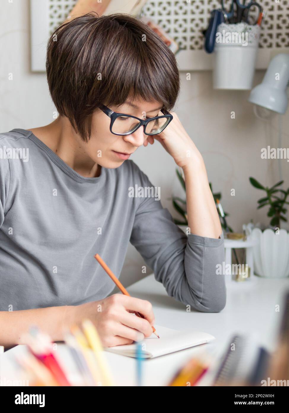 Donna con taglio corto dei capelli sta disegnando nel taccuino. Hobby calmante, tempo libero antistress. Artista al lavoro. Ambiente di lavoro accogliente. Foto Stock