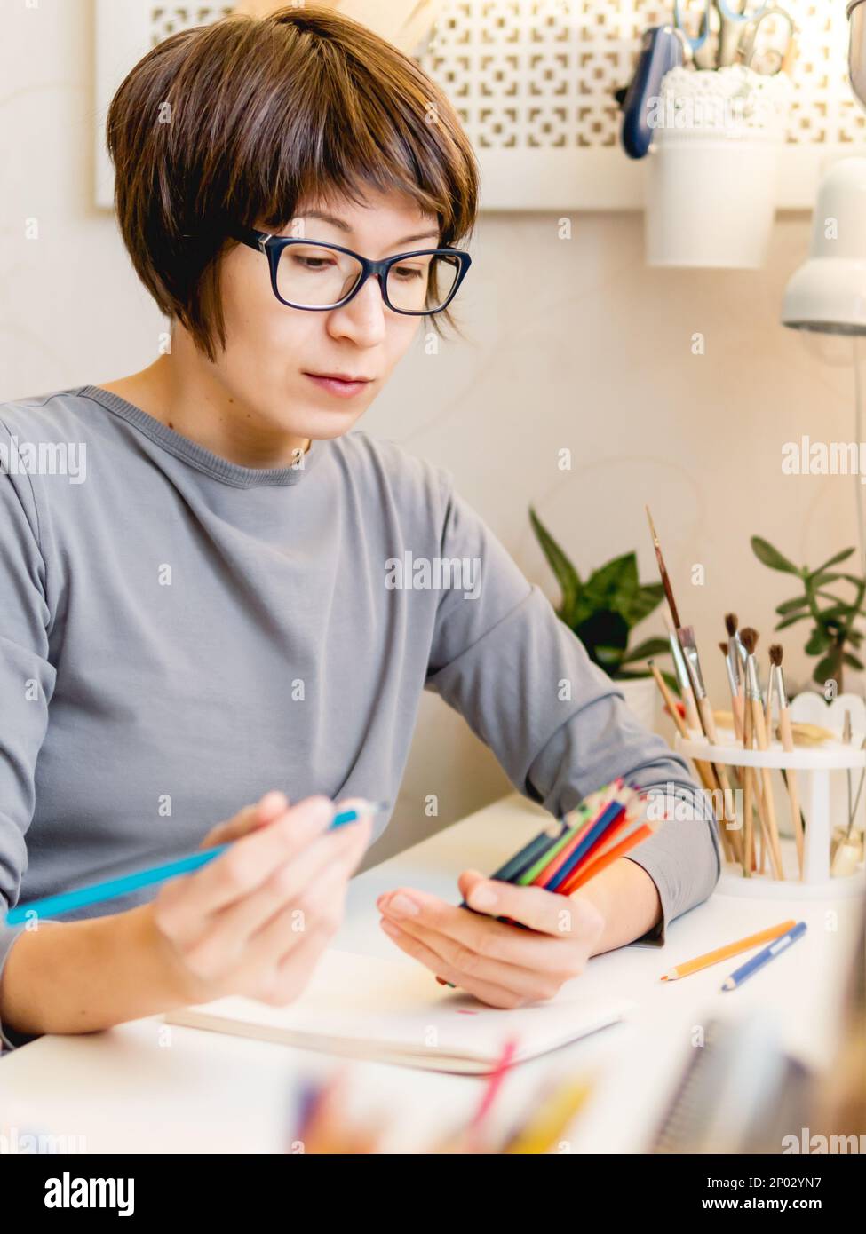 Donna con taglio corto dei capelli sta disegnando nel taccuino. Hobby calmante, tempo libero antistress. Artista al lavoro. Ambiente di lavoro accogliente. Foto Stock