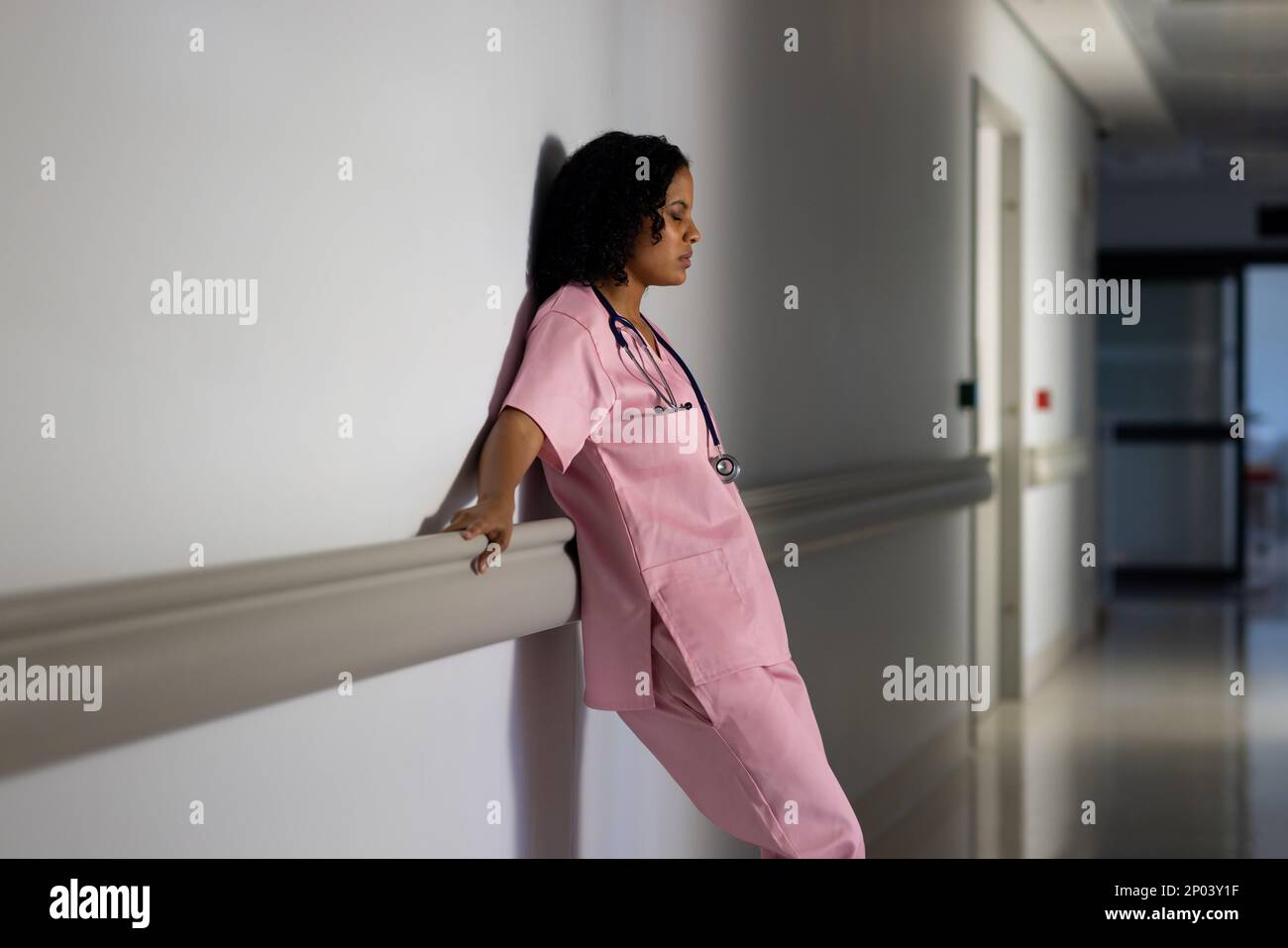 Medico di sesso femminile biraciale esausto con uno stetoscopio appoggiato alla parete in ospedale Foto Stock