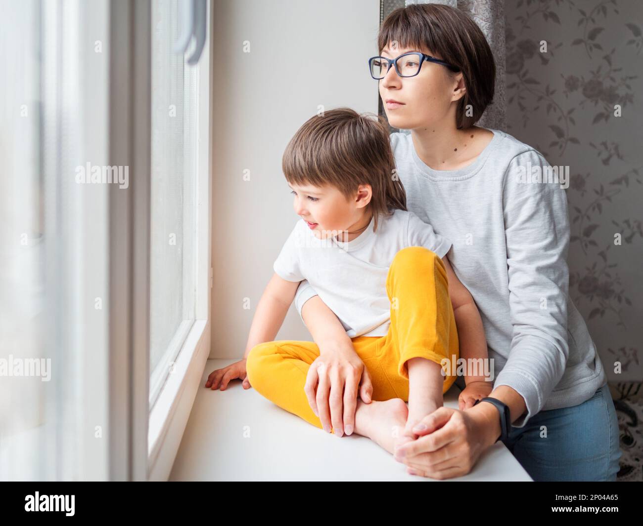 Donna triste guarda la finestra. Soggiorno forzato a casa a causa di coronavirus COVID-19 quarantena pandemia. Madre e figlio. Mamma e bambino. Foto Stock