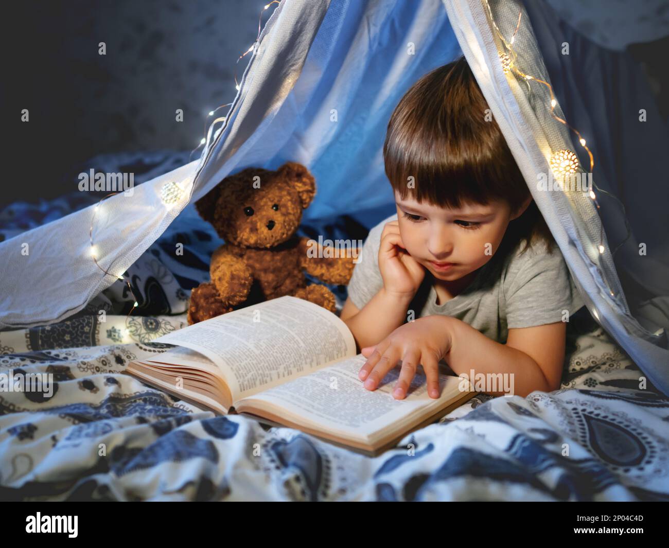 Il ragazzino legge il libro. Il bambino gioca in tenda fatta di lenzuola di lino con lampadine sul letto. Serata accogliente con il libro preferito. Foto Stock