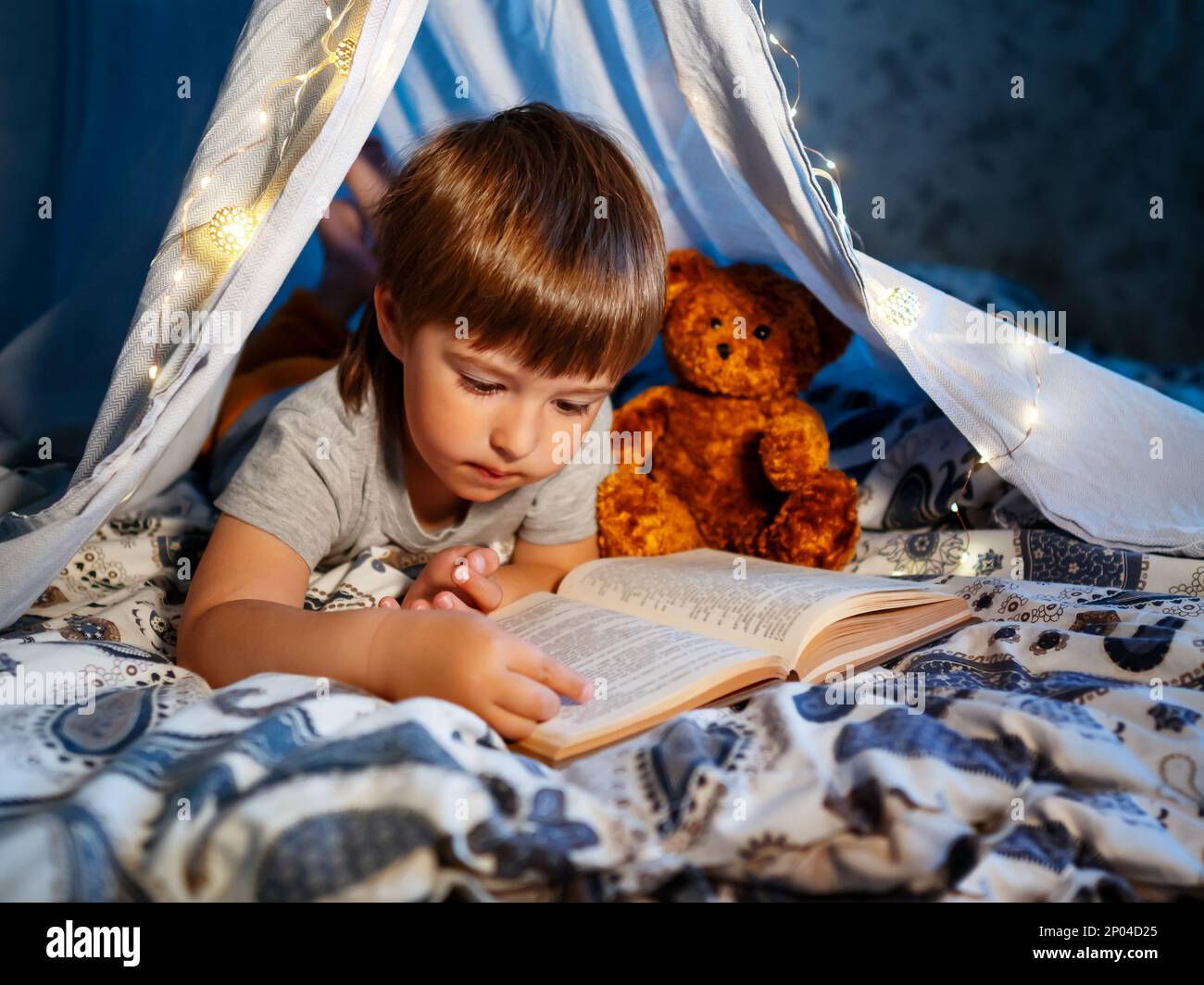 Il ragazzino legge il libro con il suo orsacchiotto. Il bambino gioca in tenda fatta di lenzuola di lino sul letto. Serata accogliente con il libro preferito. Foto Stock