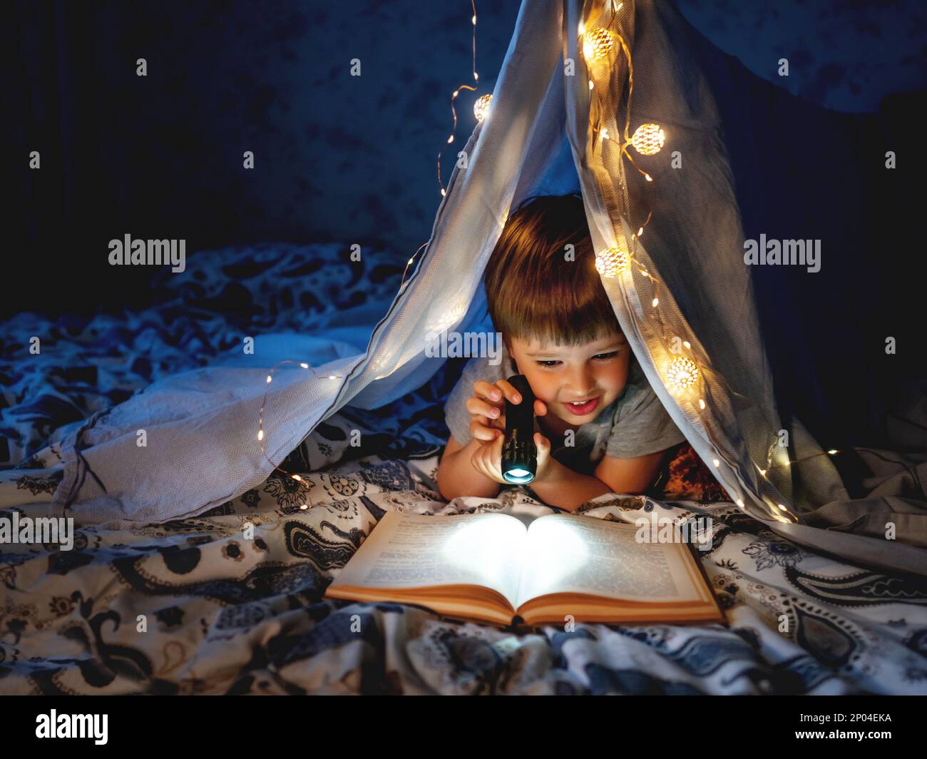 Il bambino legge con una luce tascabile. Il bambino gioca in tenda fatta di lenzuola sul letto. Serata accogliente con il libro preferito. Foto Stock
