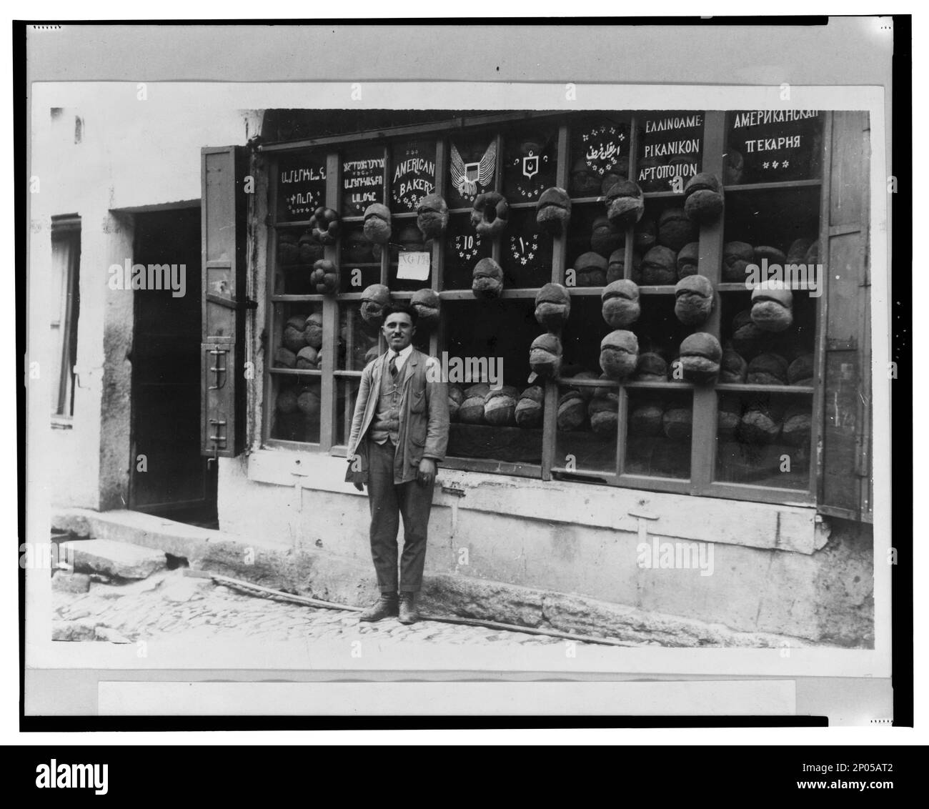Baker in piedi di fronte alla "panetteria americana" che mostra segni in armeno, Ladino (in caratteri ebraici), inglese, ottomano turco, greco e russo con campioni di pane attaccato ai mullioni, Ortaky̲, Istanbul, Turchia / foto di cdm. Frank and Frances Carpenter Collection , pubblicato in: 'Il mondo e le sue culture' capitolo del ebook grandi fotografie dalla Biblioteca del Congresso, 2013, panetterie,Turchia,Istanbul,1910-1920, pane,Turchia,Istanbul,1910-1920, segni (Avvisi),Turchia,Istanbul,1910-1920. Foto Stock