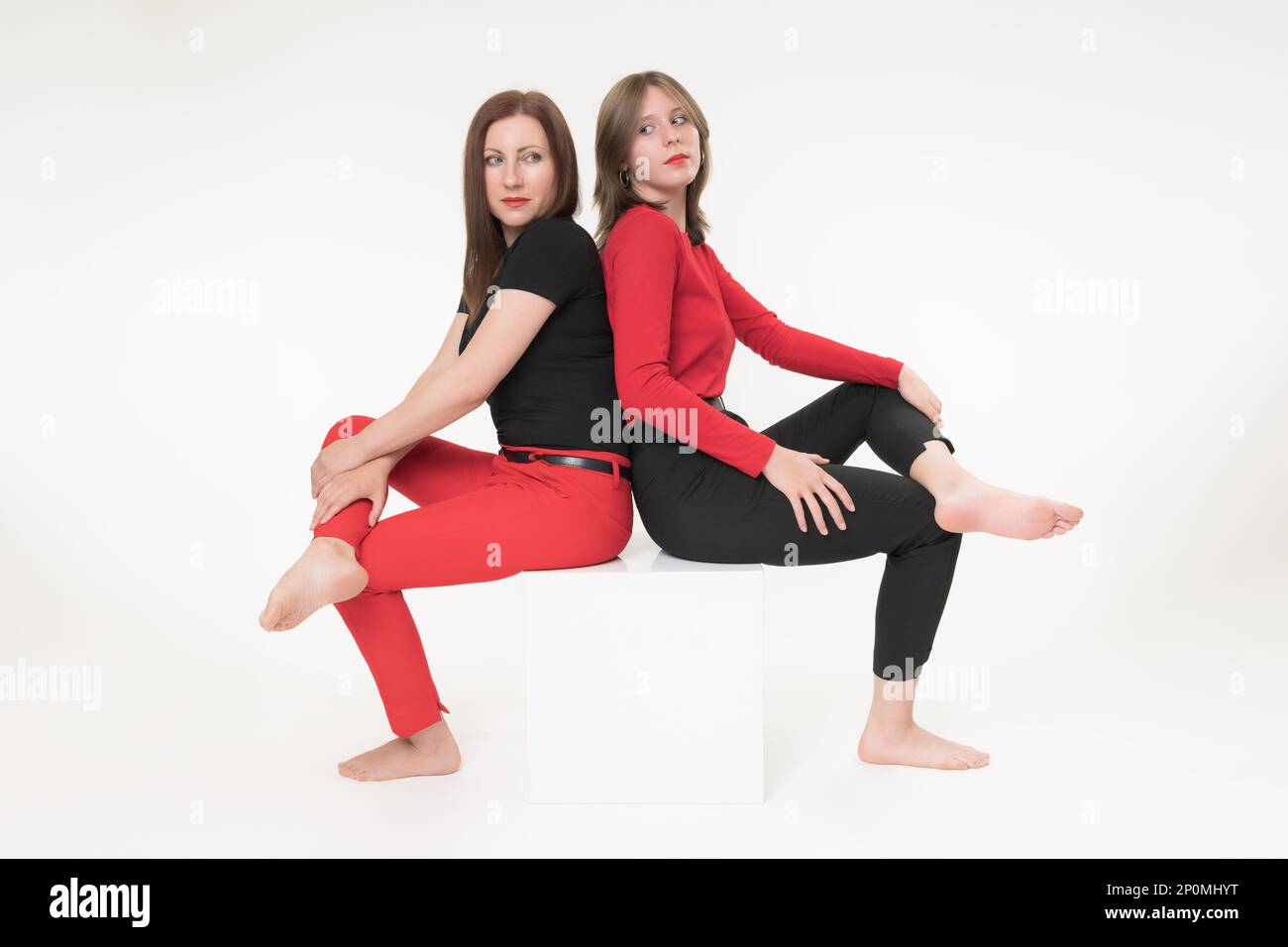 Donna mamma e figlia seduta con le spalle l'una all'altra su cubo su sfondo bianco. Entrambe le donne hanno attraversato le gambe e guardando seriamente Foto Stock