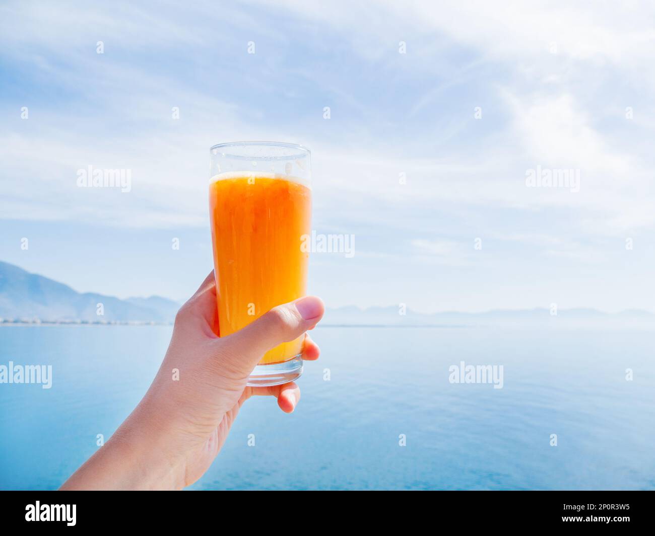 A mano con il succo fresco e gustoso di arance mature spremute nel bicchiere. Mare calmo con montagne all'orizzonte. Kemer, Turchia. Foto Stock