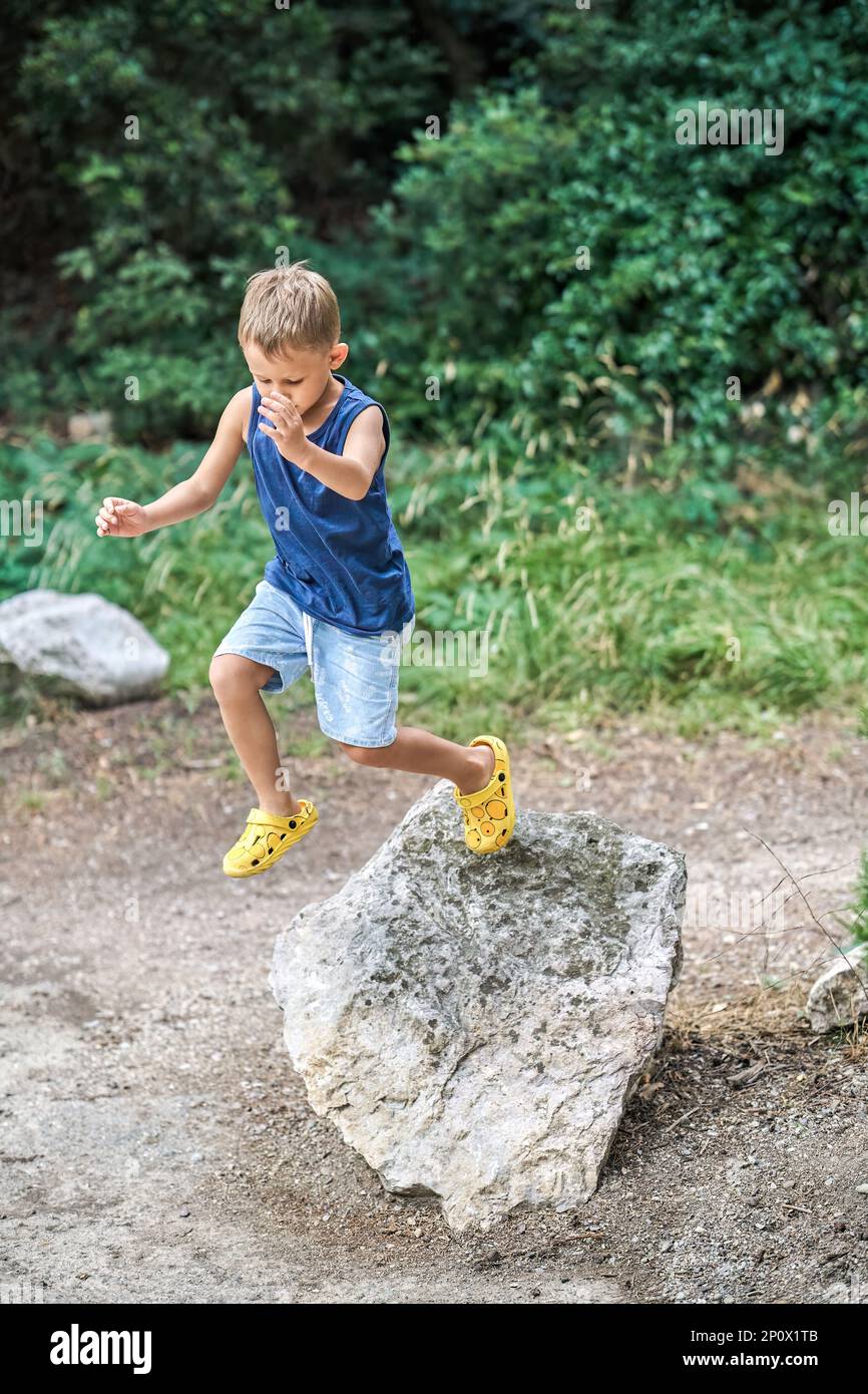 Il ragazzino attivo salta giù dalla grande roccia nel verde del parco estivo. Carino bambino preschooler in abiti casual gioca nel parco naturale selvaggio durante la passeggiata Foto Stock