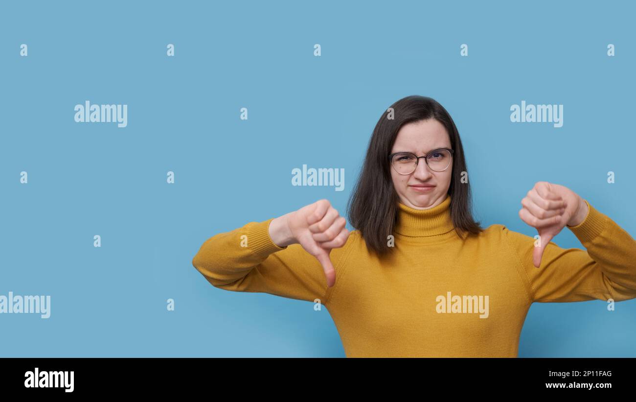 Un ritratto di una studentessa emotiva insoddisfatta in occhiali e un maglione giallo che punta il dito verso il basso su uno sfondo blu. Emozione negativa. Foto Stock