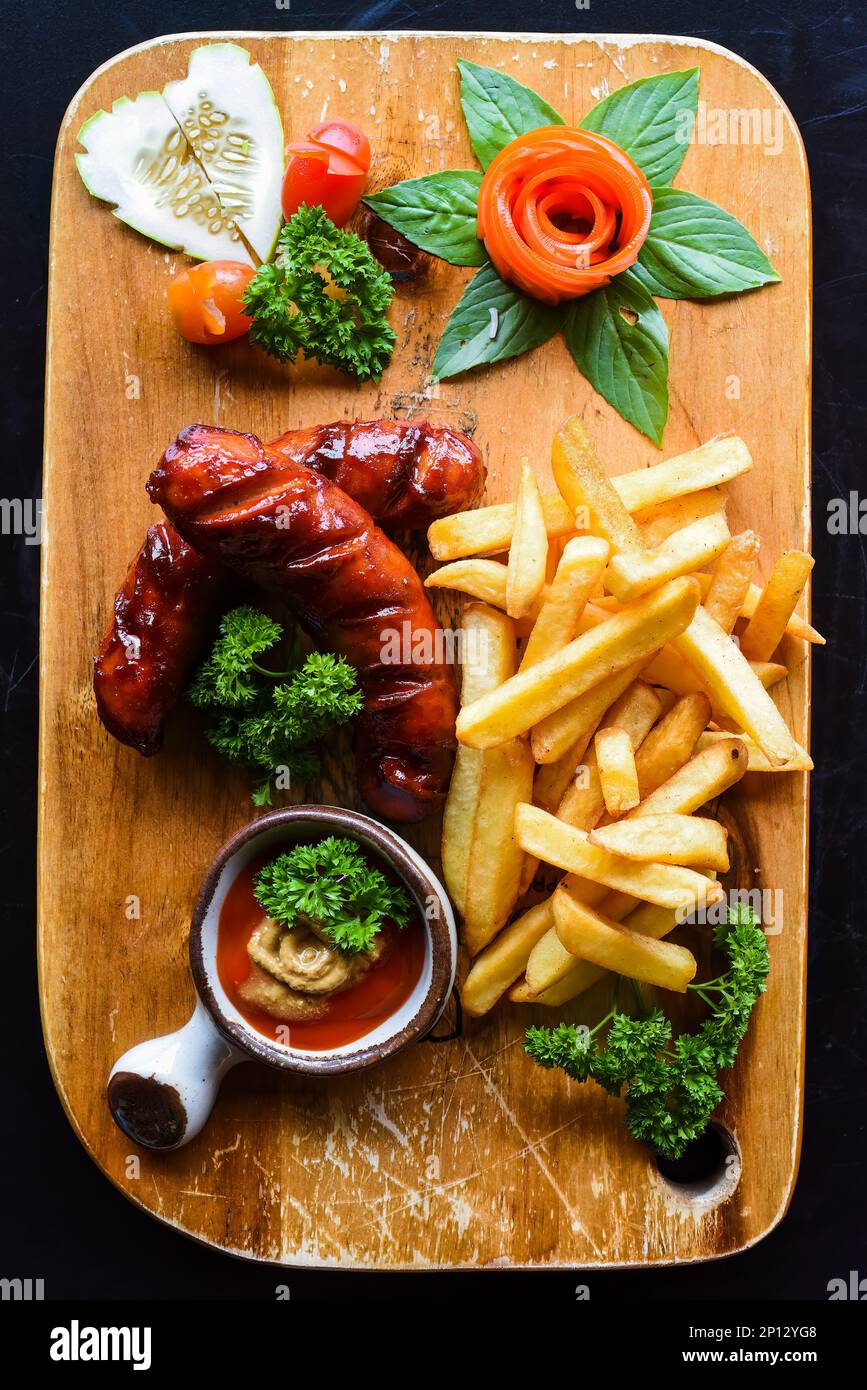 Salsicce fritte con patatine fritte e salsa piccante su asse di legno isolato su sfondo nero vista dall'alto Foto Stock