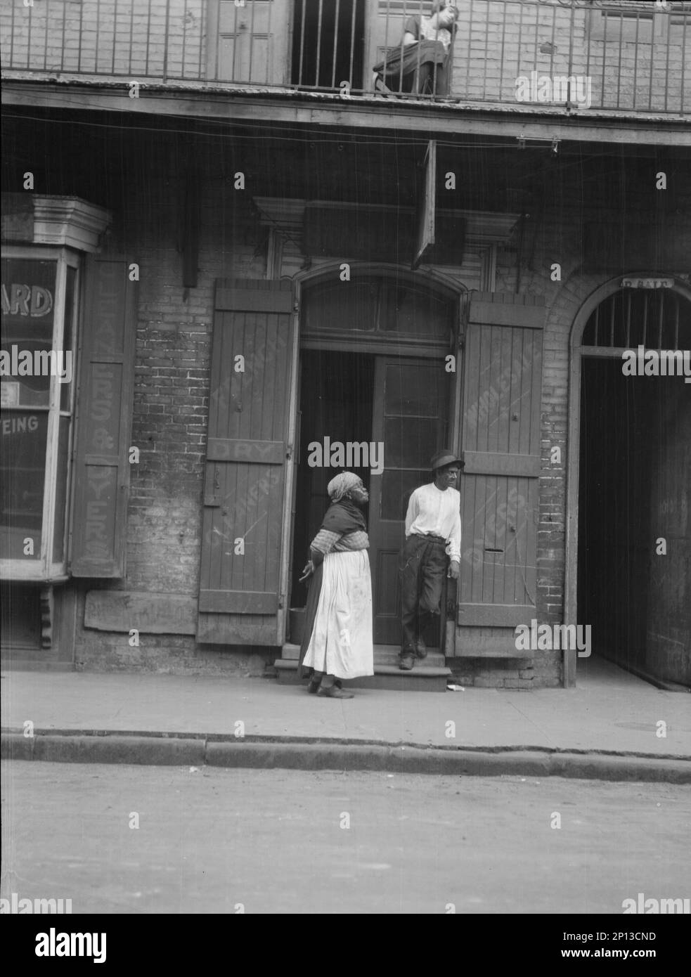 Vista dall'altra parte della strada di un uomo e di una donna in piedi accanto a una porta e di una donna seduta sul balcone sopra, New, tra il 1920 e il 1926. Foto Stock