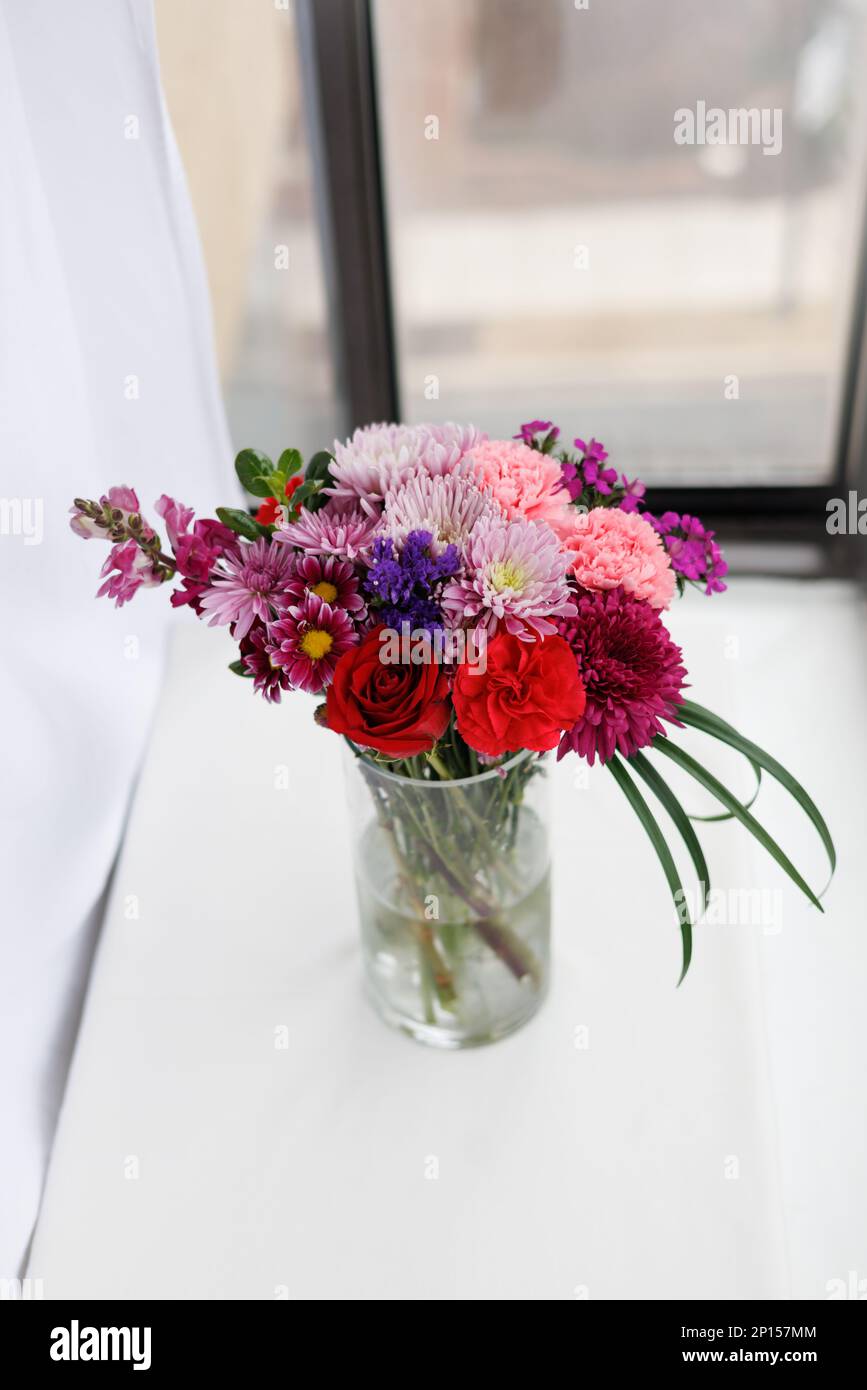 Fiori rossi, rosa, viola disposti in un vaso di vetro vicino alla finestra Foto Stock