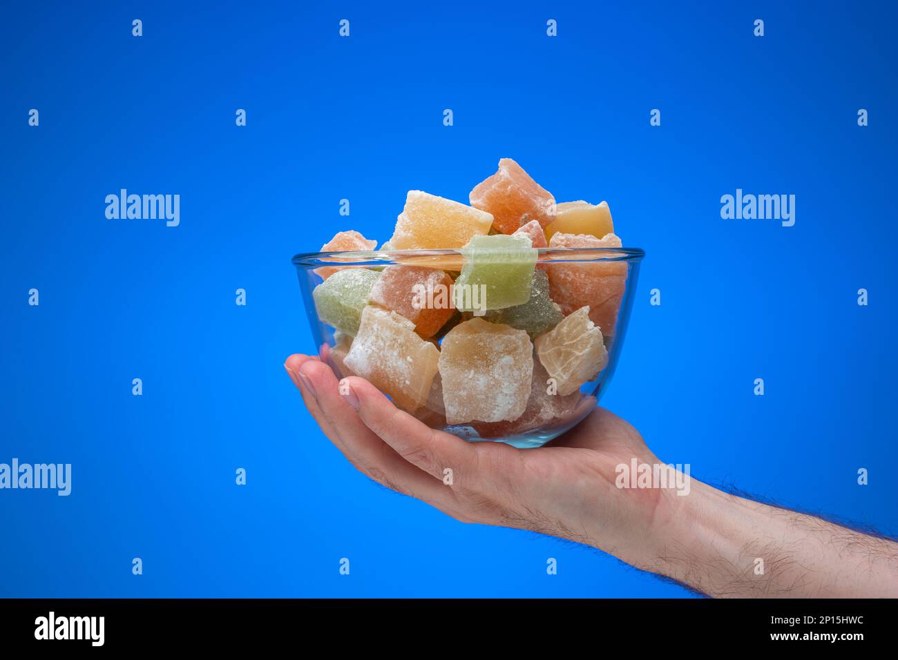 Cubetti di delizia turca colorata in una ciotola di vetro tenuta in mano maschile. Primo piano ripresa in studio, isolata su sfondo blu. Foto Stock