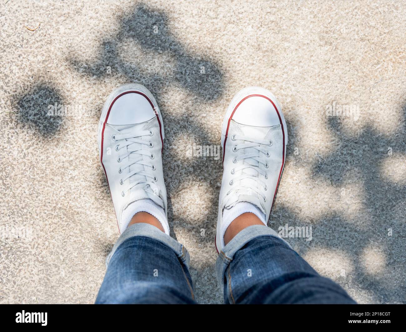 Piedi donna in sneakers bianche su pavimento in pietra. Vista dall'alto su abiti e scarpe informali alla luce del sole. Atmosfera estiva. Moda urbana moderna. Foto Stock