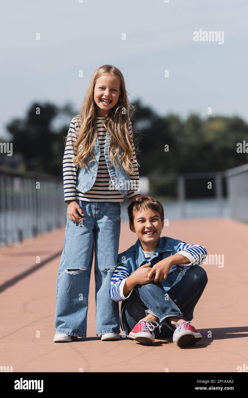 lunghezza intera di ragazza sorridente in vestito di denim in piedi vicino al ragazzo felice di presen sul argine del fiume, immagine di scorta Foto Stock