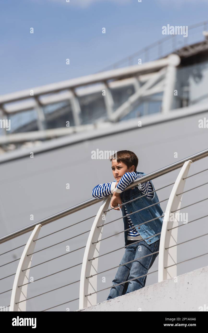 vista ad angolo basso del ragazzo in denim gilet e jeans appoggiati su recinzione metallica sul terrapieno, immagine stock Foto Stock