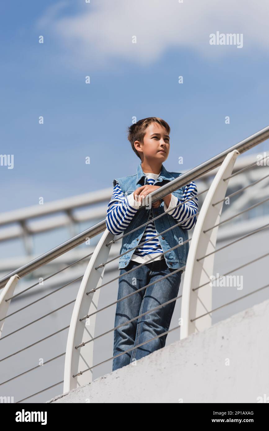 vista ad angolo basso del ragazzo in jeans outfit alla moda che posa vicino a recinzione metallica sul terrapieno, immagine stock Foto Stock
