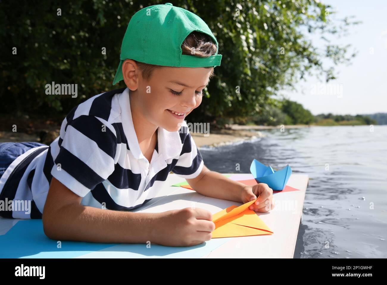 Carino ragazzino che fa barche di carta sul molo vicino al fiume Foto Stock