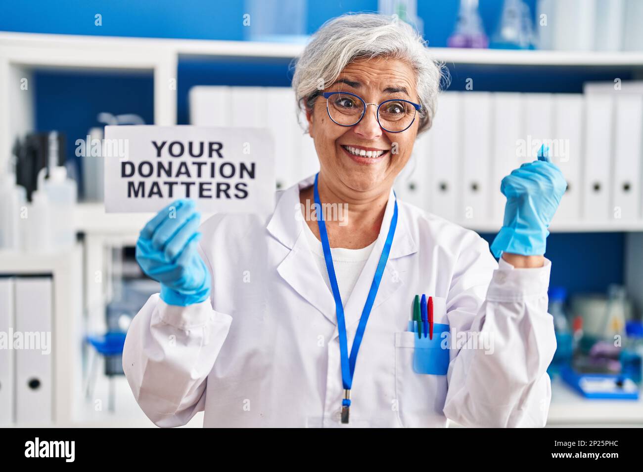 Donna di mezza età con i capelli grigi che lavorano al laboratorio di scienziato che tiene la vostra donazione importa banner urlando fiero, celebrando la vittoria e il successo Foto Stock