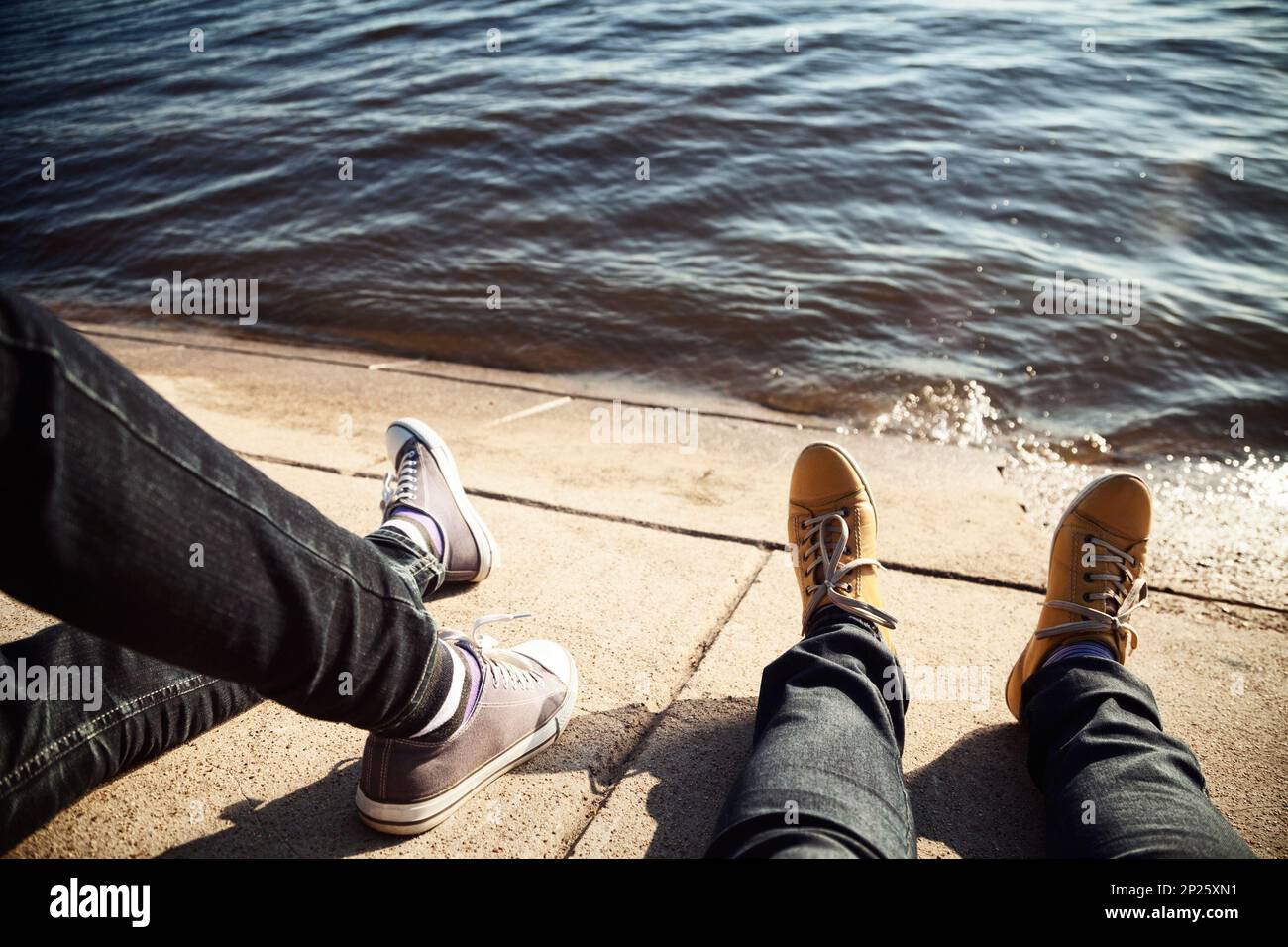 Gambe di due amici appendere fuori insieme vicino all'acqua. 2 giovani seduti e rilassarvi su una riva di un fiume Foto Stock