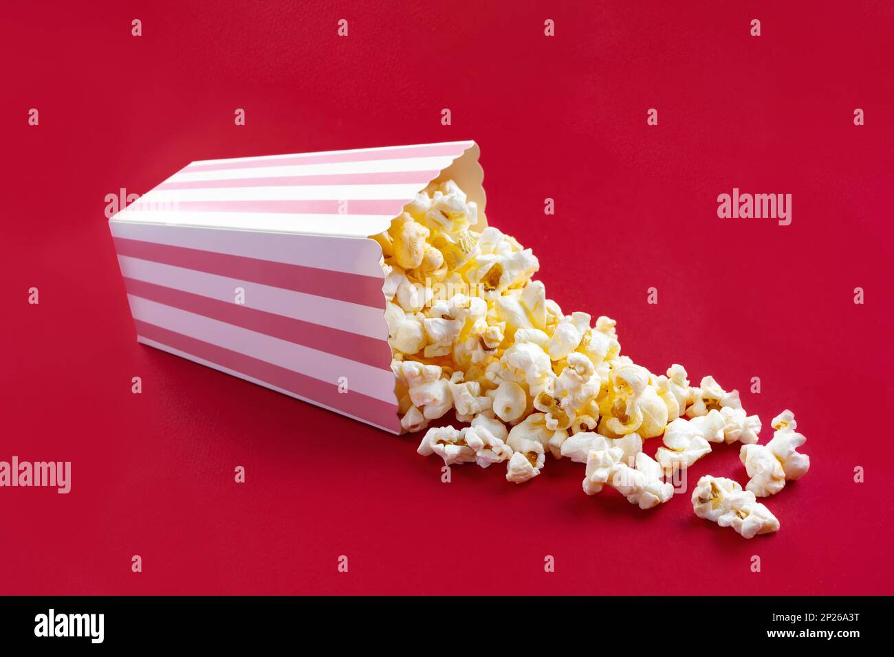 Gustoso popcorn al formaggio che cade da un secchio di cartone a strisce rosa, isolato su sfondo rosso. Dispersione di grani di popcorn. Film, cinema e intrattenimento Foto Stock