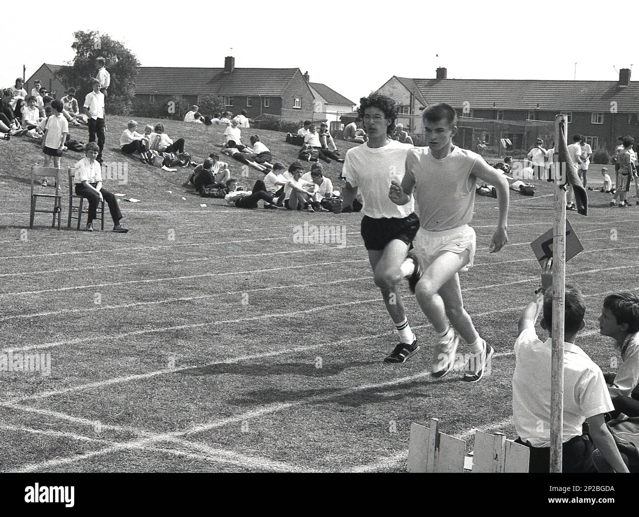 1989, una giornata di sport in una scuola secondaria, due ragazzi in gara in una corsa in erba di un campo sportivo, Sleaford, Lincs, Inghilterra, Regno Unito. Un scolaro seduto sull'erba tiene un numero di giro. Foto Stock