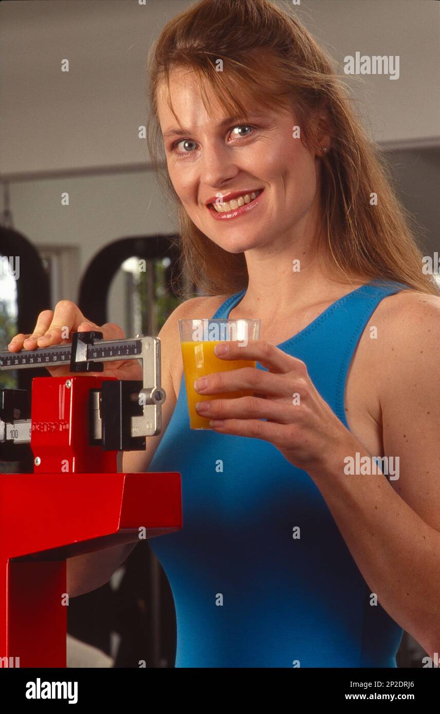 Giovane donna in palestra, in pausa dal suo lavoro per salire sulla bilancia e pesarsi mentre tiene un bicchiere di succo d'arancia Foto Stock