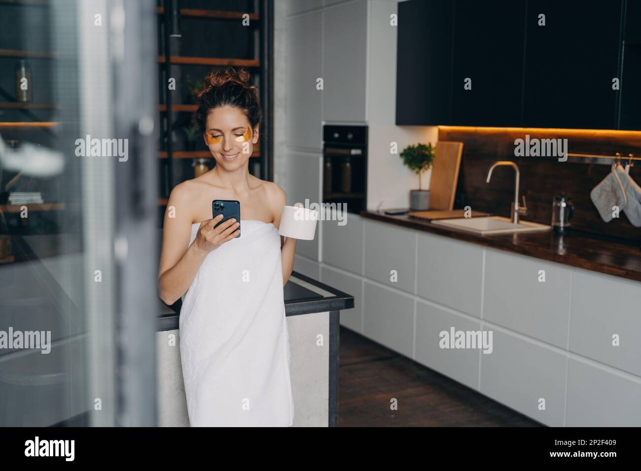 Bella donna con macchie sotto gli occhi, utilizzando lo smartphone in cucina. Ragazza sorridente avvolta in asciugamano bere caffè leggendo le notizie in linea sui social media Foto Stock