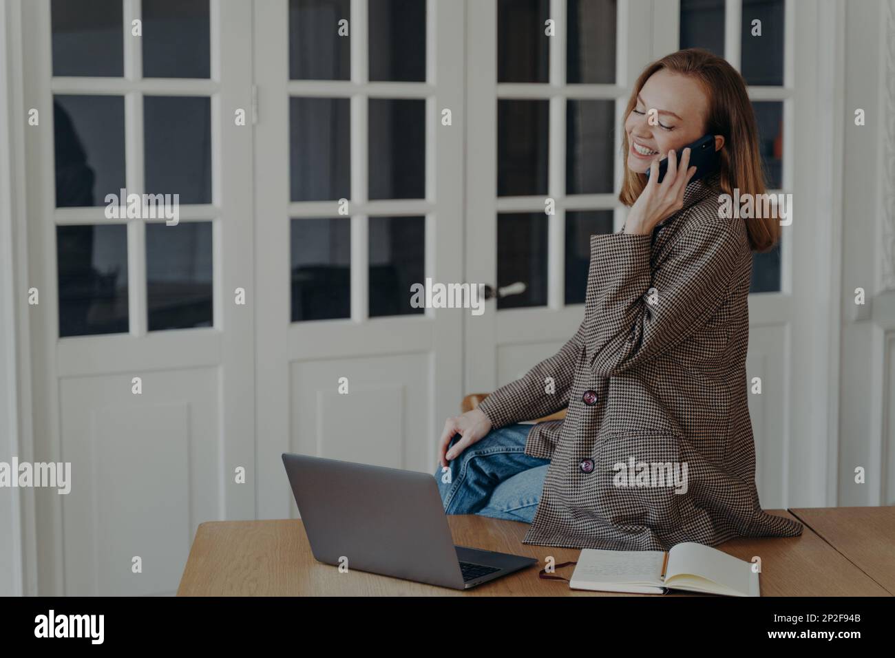 Una donna sorridente, freelancer, risponde alle chiamate mobili, seduto sulla scrivania con un laptop che parla per telefono. Felice giovane donna dipendente siede al tavolo ufficio cha Foto Stock