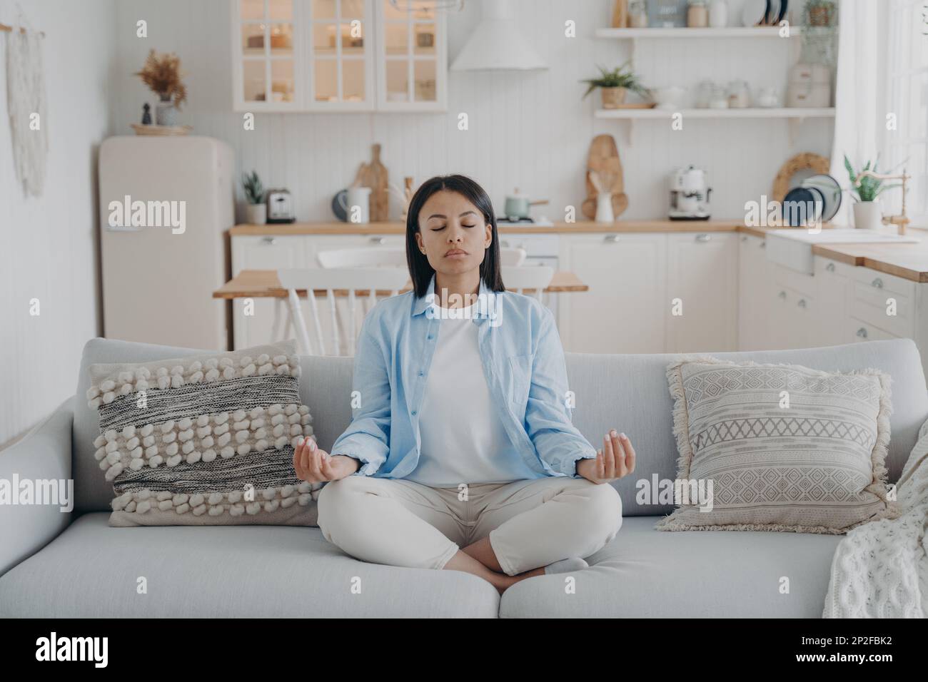 Donna serena che pratica yoga e meditazione tenendo le mani nel gesto mudra, seduto comodo divano a casa. Calma giovane donna che si rilassa sul divano. Benessere, Foto Stock