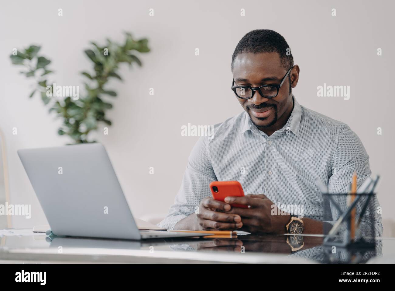 Felice uomo d'affari afro in occhiali sta testando su smartphone. Il freelance sta lavorando online sul pc e sta conversando con il cliente nell'ufficio di casa. REM Foto Stock