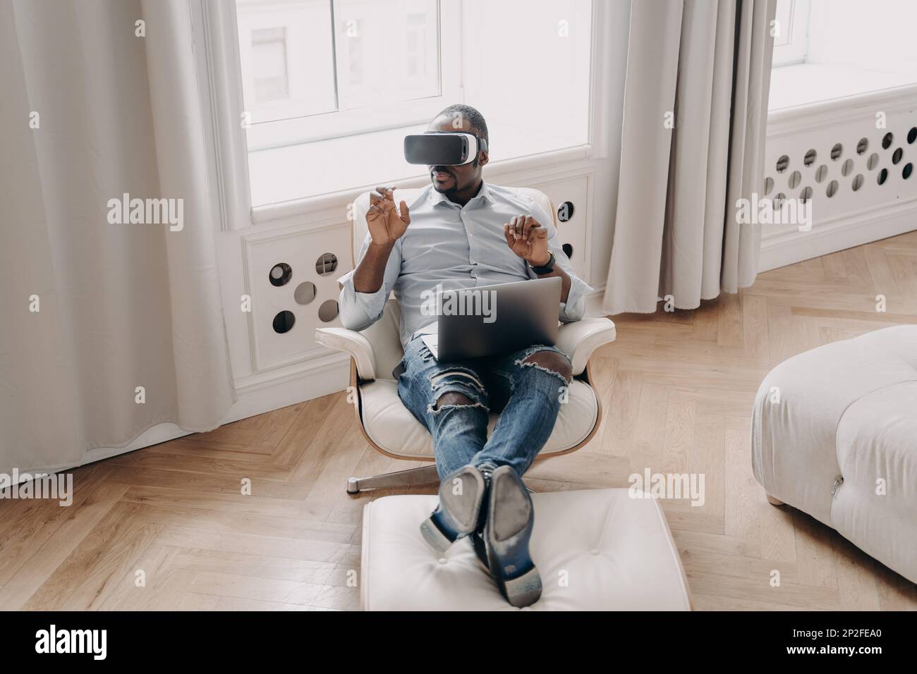 Uomo afroamericano in occhiali VR che lavorano in realtà aumentata, seduto in poltrona con il laptop a casa, puntando l'oggetto di realtà virtuale, interagisci Foto Stock