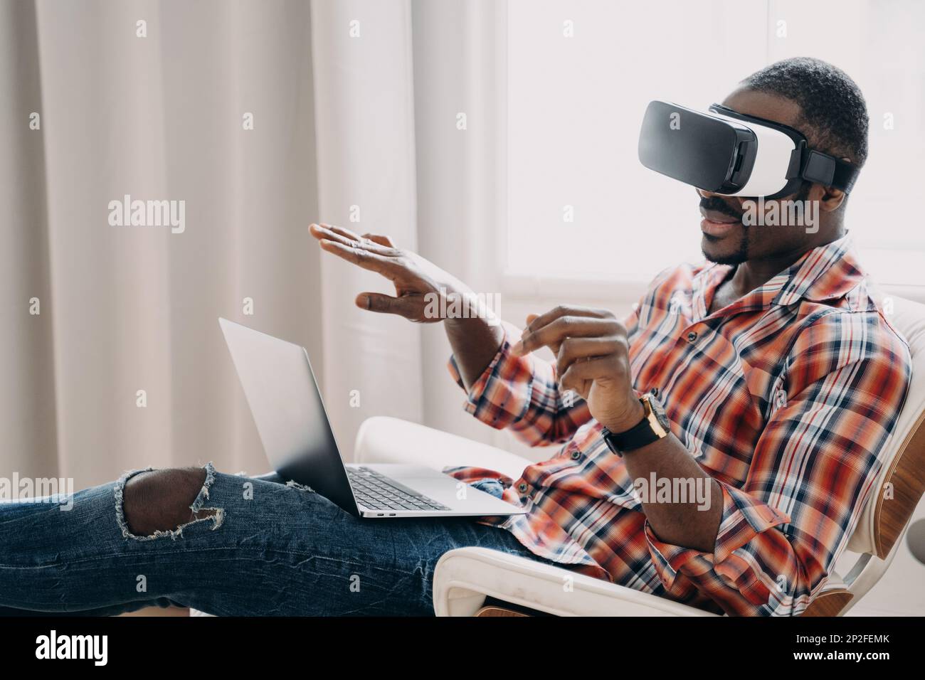 Sorridente afro-americano in occhiali VR si siede in poltrona con il laptop, testando gli occhiali per la realtà virtuale. Moderno uomo nero che guarda l'acquisto in cybersp Foto Stock