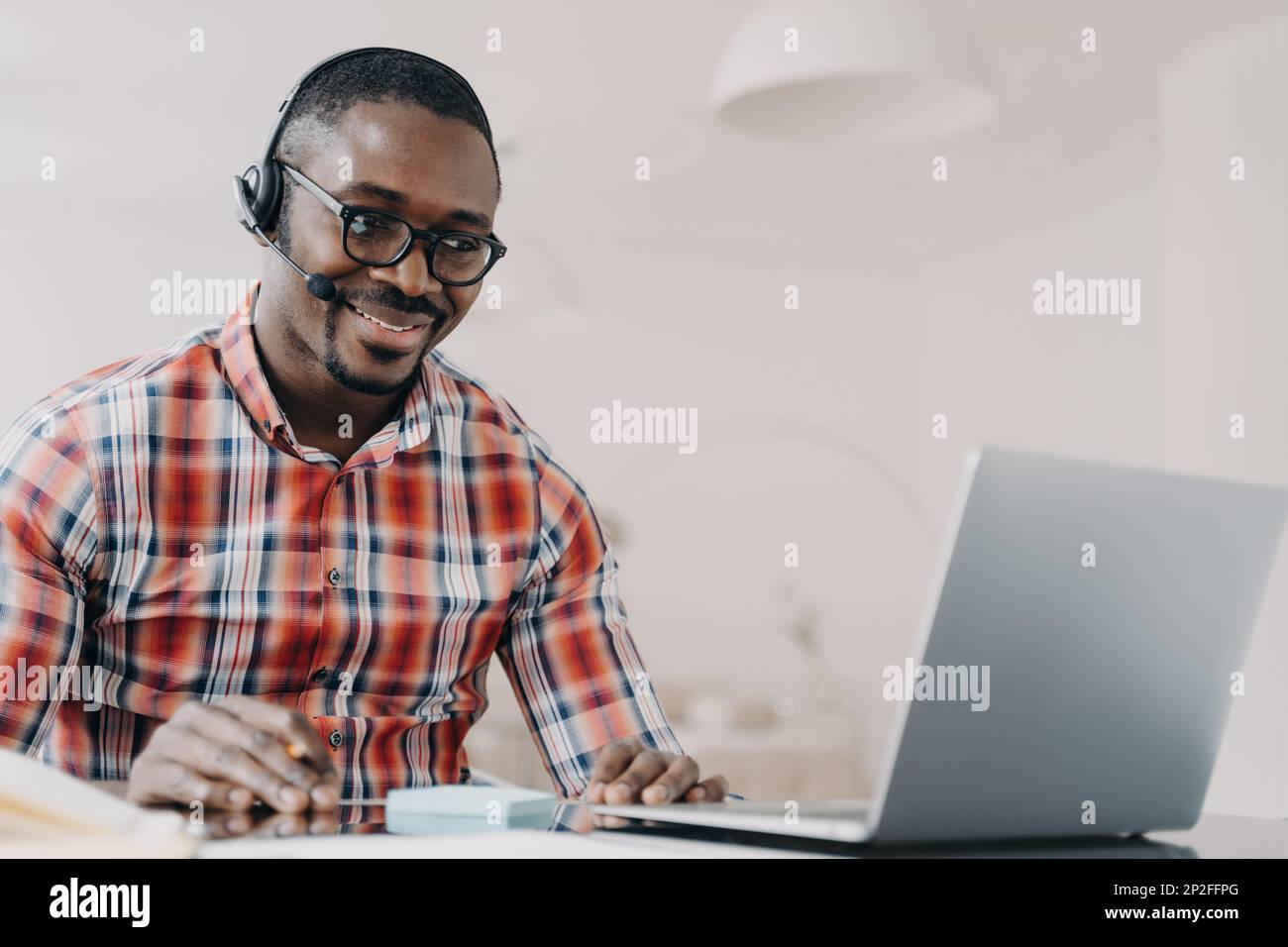Sorridente dipendente afro-americano che indossa cuffie che lavorano sul computer portatile, guarda lo schermo, risponde a una videochiamata, comunica con il cliente. Ragazzo nero Foto Stock