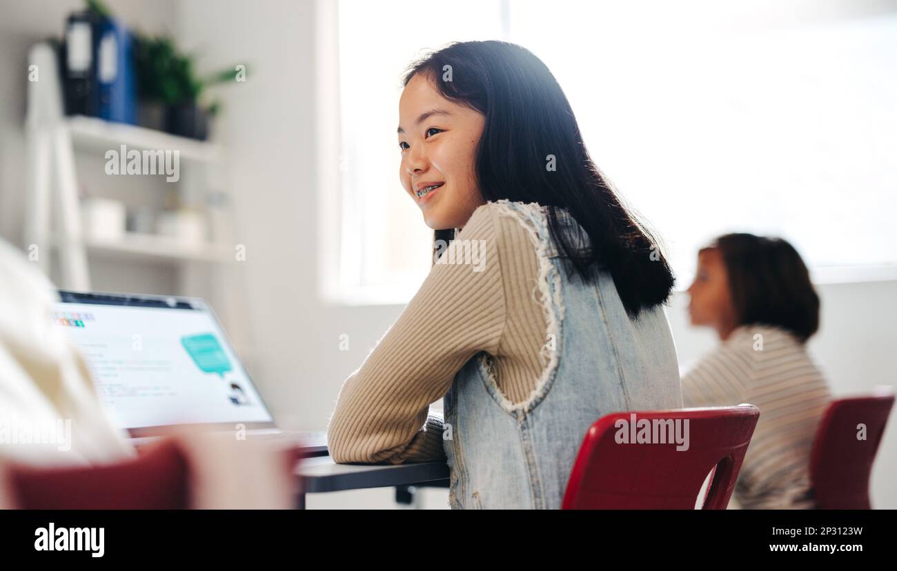 Giovane ragazza asiatica seduta in una classe di codifica con un computer portatile davanti a lei e un vivo interesse per il suo volto. Desideroso di padroneggiare le sue abilità di programmazione, lei Foto Stock