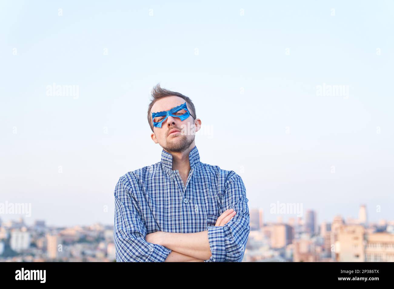 Uomo caucasico in abiti da sonno e maschera blu supereroe in piedi all'aperto con faccia seria. Vista urbana sullo sfondo. Immagini di alta qualità Foto Stock