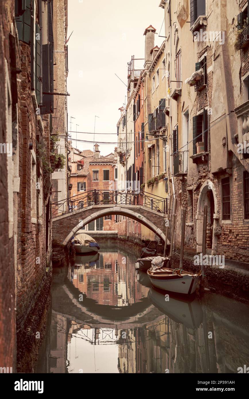 Un ponte pedonale ad arco e vecchie case sul canale si riflettono in uno stretto canale di Venezia accanto a barche ormeggiate, Italia, in una composizione verticale vintage. Foto Stock