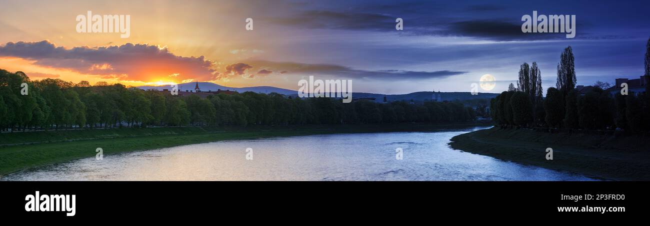 il giorno e la notte cambiano sopra la città di uzhhorod. paesaggio urbano con fiume con peccato dietro la montagna e la luna sopra gli alberi al crepuscolo Foto Stock