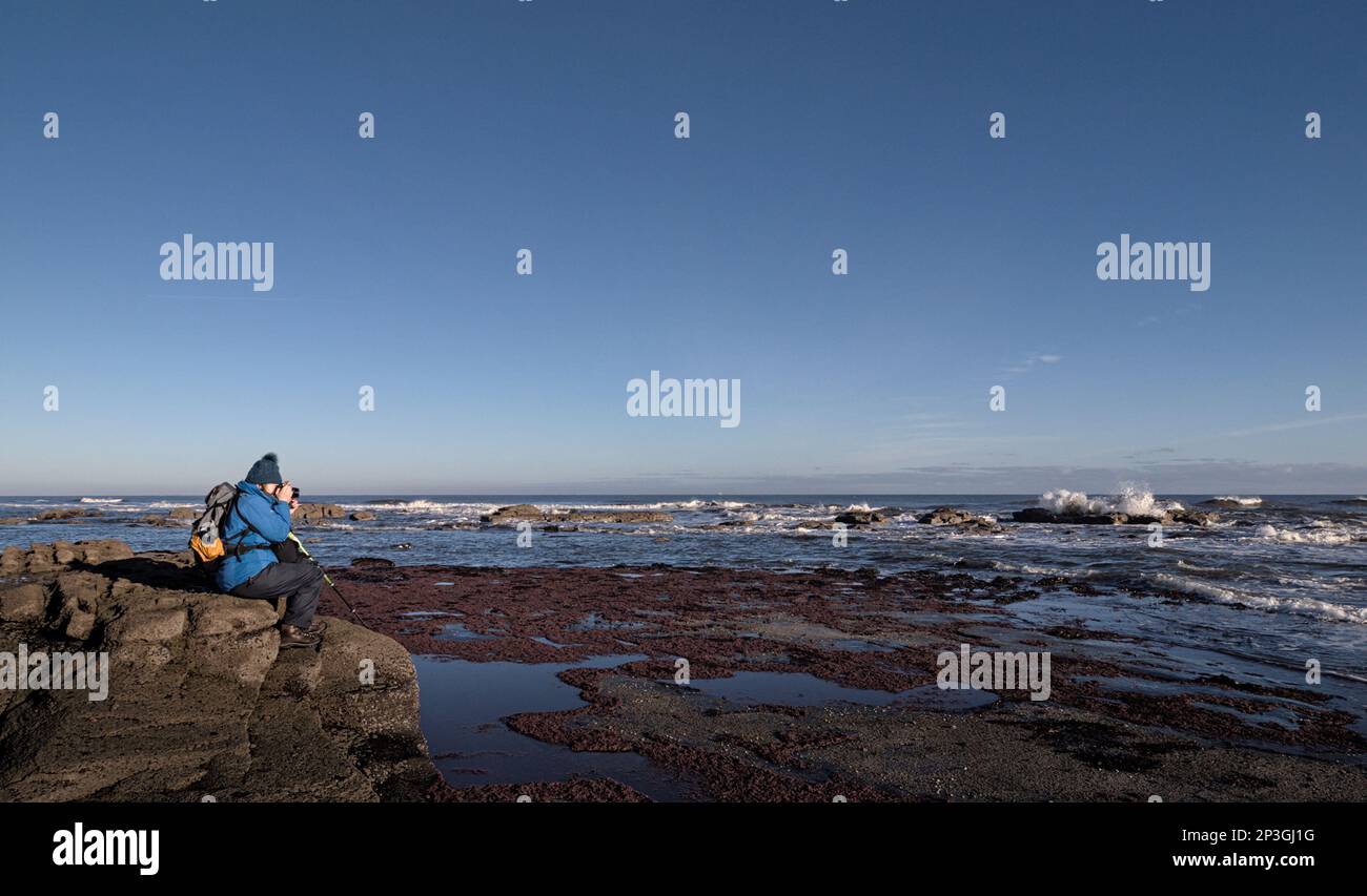 Una fotografa femminile scatta foto delle onde a Saltwick Bay vicino Whitby, North Yorkshire Foto Stock