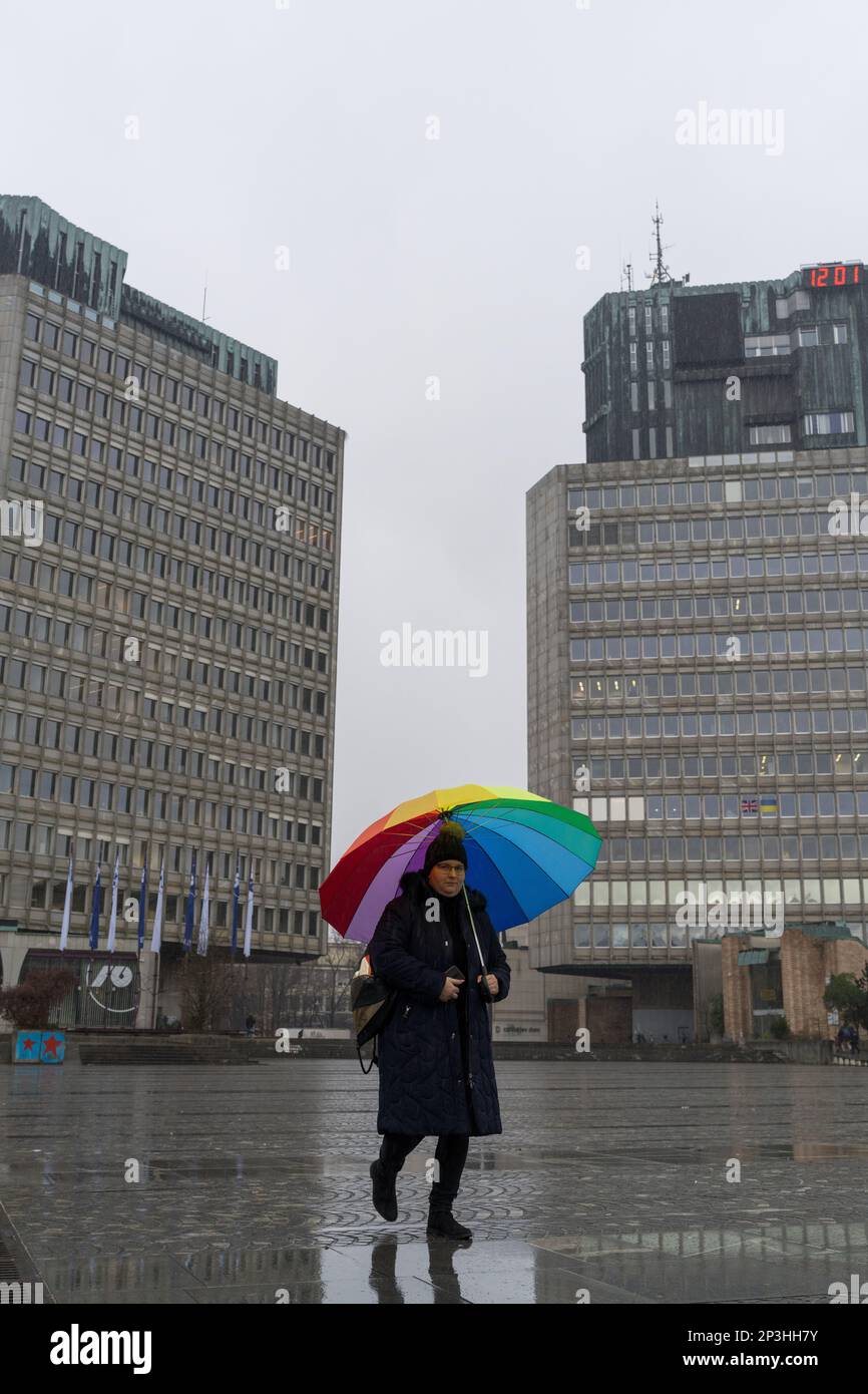 12 26 2022, Lubiana, Slovenia: Una donna con un ombrello colorato che cammina giù Piazza della Repubblica / Trg Repubblica incorniciata da Lubijanska Banka e TR3 to Foto Stock