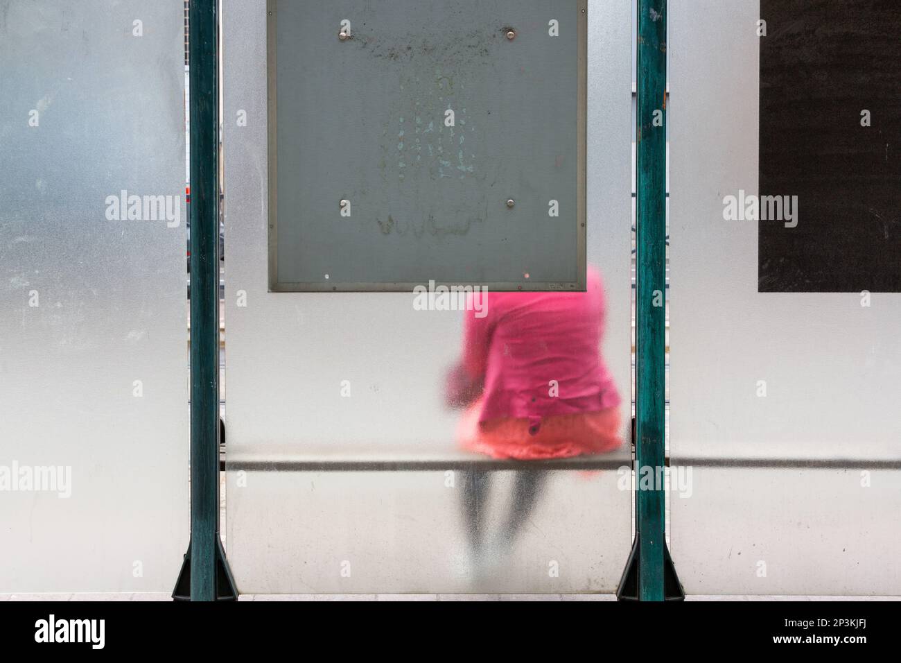 Signora in un cappotto rosa seduto in un rifugio di autobus con pareti di vetro smerigliato. Bruxelles. Foto Stock