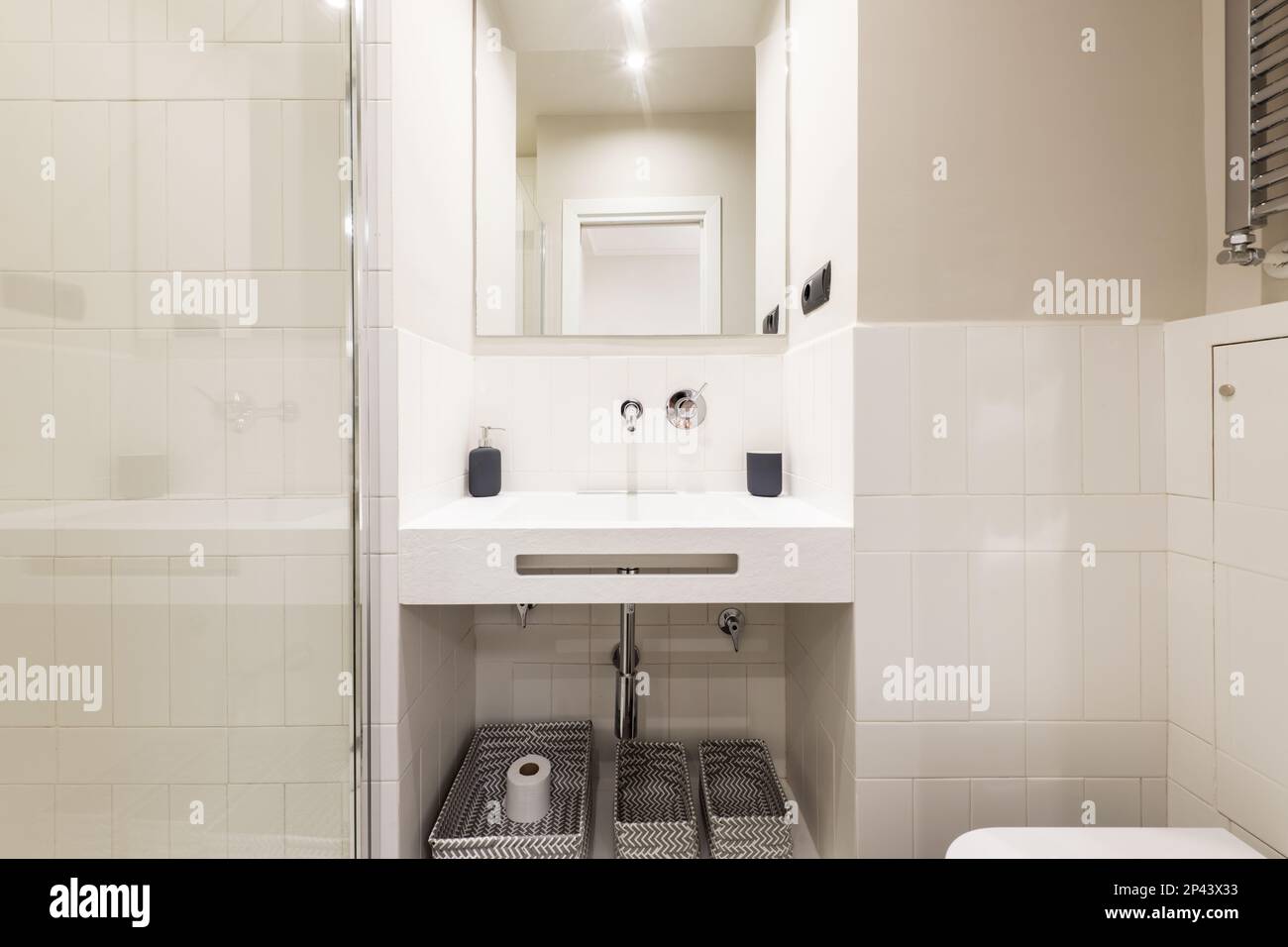 Bagno con lavabo dal design inusuale, rubinetto cromato e scaldasalviette, specchio senza cornice, cestini in tessuto sotto il lavabo e divisorio in vetro temprato Foto Stock