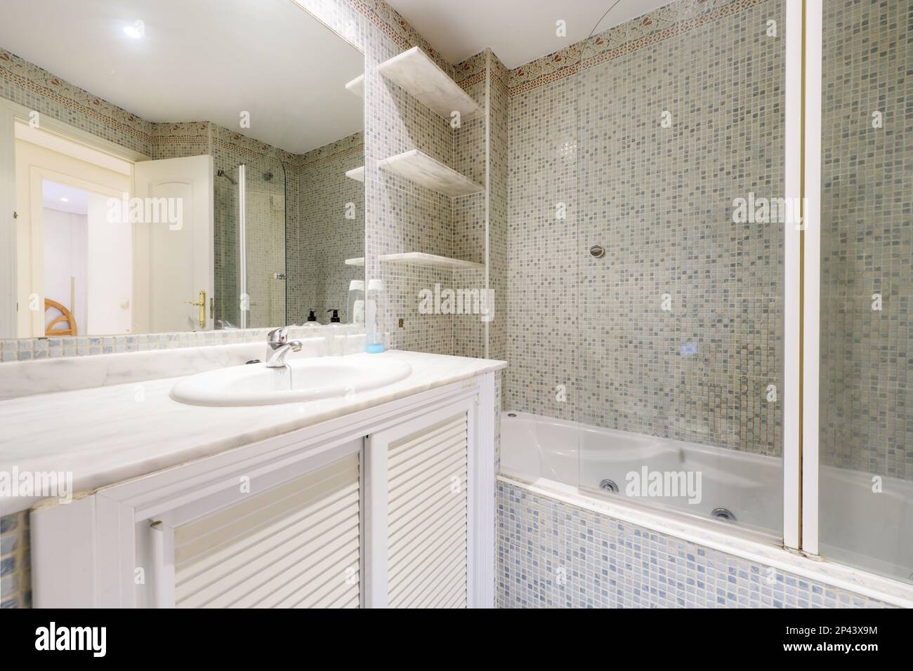 Bagno con lavabo in porcellana bianca, piano in marmo, rubinetti cromati, specchio senza cornice, ripiani in marmo e pareti piastrellate con gresite Foto Stock