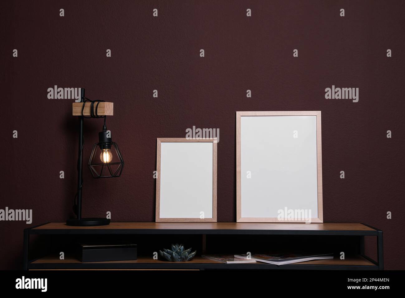 Cornici vuote e lampada elegante su tavolo di legno vicino a parete marrone. Modello per il design Foto Stock